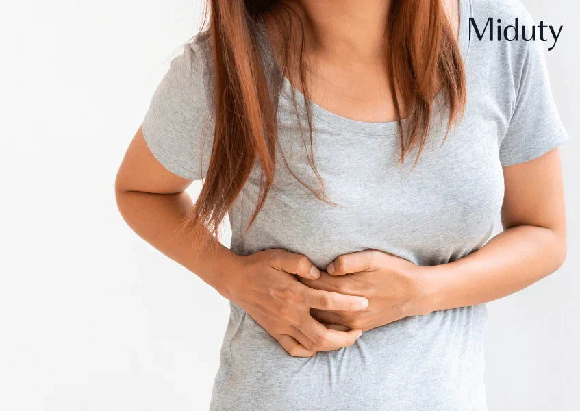 Person with brown hair in a gray shirt, holding their stomach in discomfort