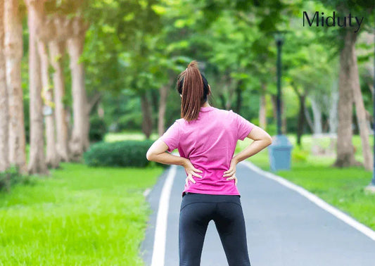 Person in athletic wear standing on a path, holding lower back, surrounded by trees