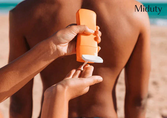 Person applying sunscreen from an orange bottle onto another person's back at the beach